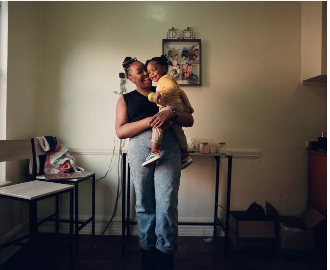 A mother holding her child in a sparse apartment, framed photos on the wall